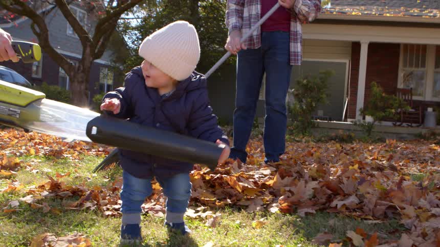 A family takes a break from raking to play in autumn leaves outside their house on a sunny day as the child plays with a leaf blower held by a parent