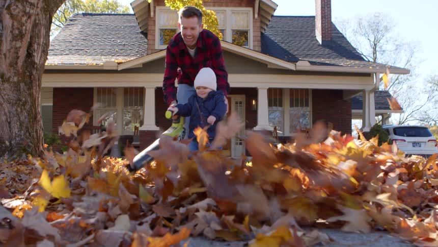 A man and child joyfully clear autumn leaves with a leaf blower in front of a suburban home