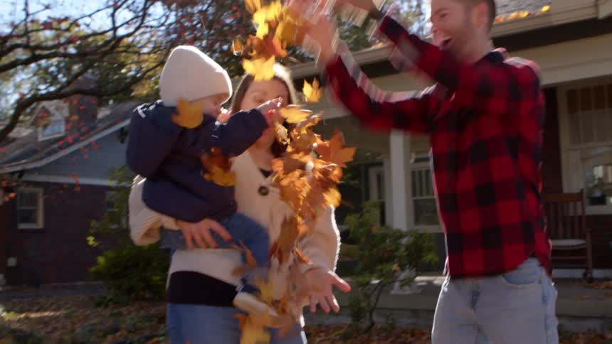 Toddler carried by his mum playfully scatters fallen leaves held up in the air by his dad as they stand outdoors during the day