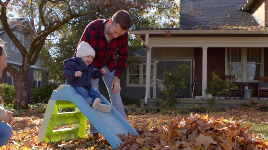 A toddler in a white hat and blue coat slides down a small green and blue slide into a pile of autumn leaves while an adult male in a plaid shirt watches and a woman crouches nearby