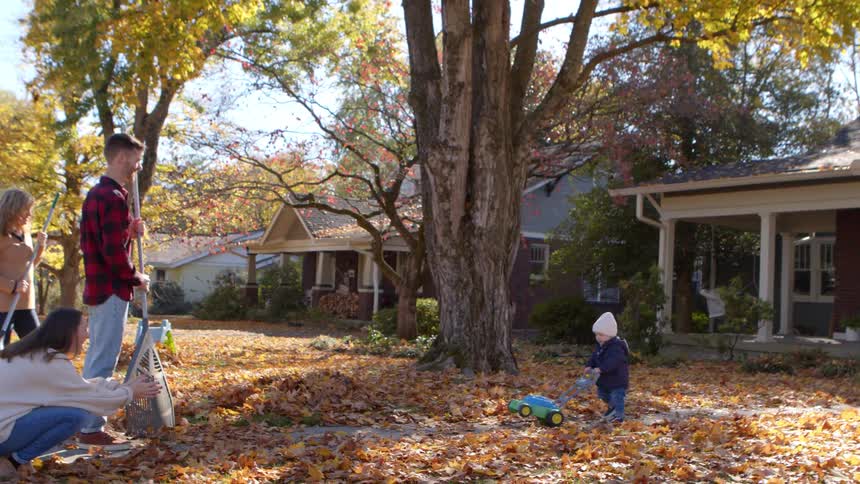 Male toddler pushes a toy mower as he walks to meet his family outdoors in front of their house during daytime