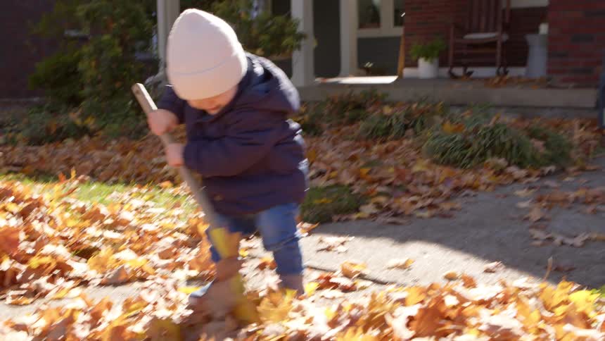 A child in a white knit hat and blue jacket rakes scattered autumn leaves on a sidewalk beside a house