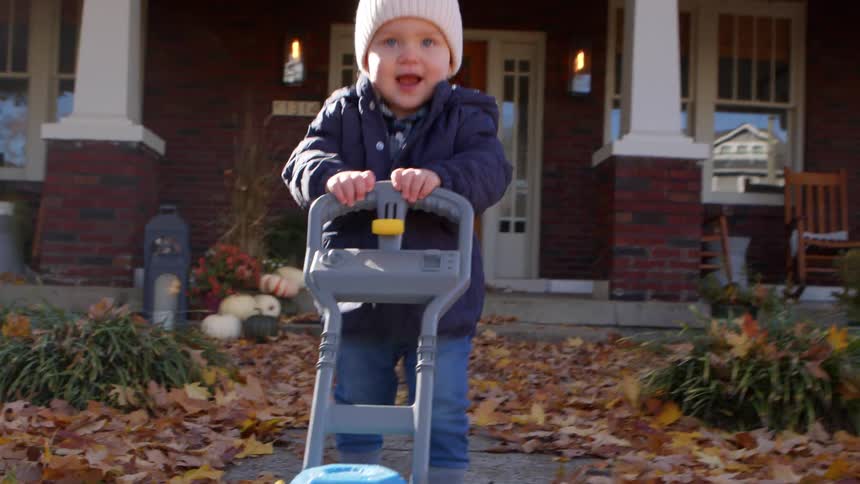 A father moves a toy lawn mower along the front yard walkway as his toddler excitedly pushes it with him at midday with the yard covered in fallen leaves