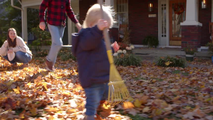 Small child accompanied by parents playing in a backyard with dry leaves fallen on the grass