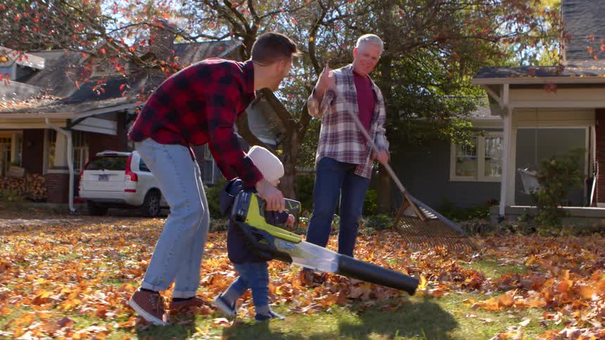 A happy family is enjoying a fun fall cleanup together using a leaf blower and rake in their front yard