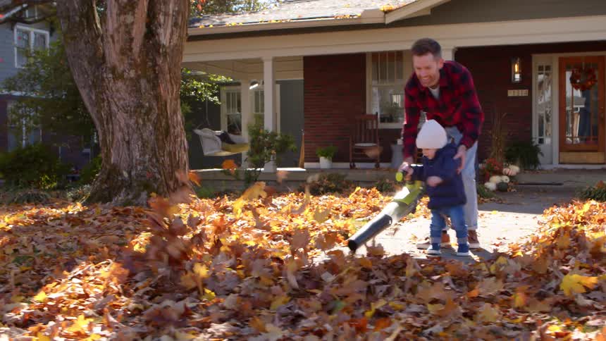 A father and child joyfully use a leaf blower together in the front yard on a sunny autumn day