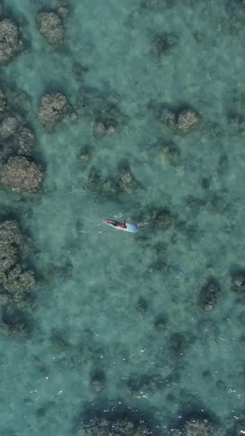 Aerial top view of a man paddling on a surfboard over clear water with large rock formations on the ocean floor below during the day