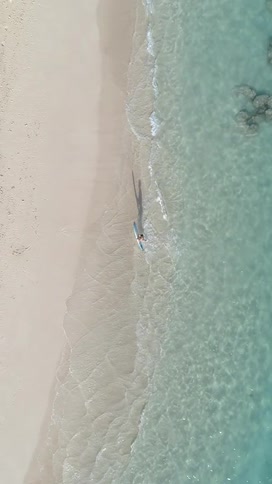 An aerial view captures a person with a blue surfboard walking on a white sand beach next to clear turquoise ocean water