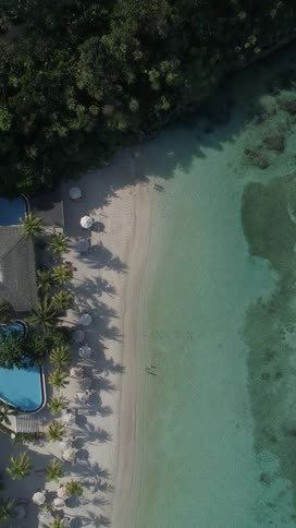 An aerial shot shows a resort building next to a sandy beach with lounge chairs and umbrellas bordering turquoise ocean water with visible coral formations
