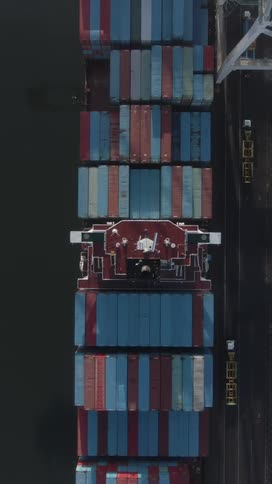 An aerial topdown view shows a large red cargo ship docked surrounded by blue and red shipping containers with dark water on the left and a concrete dock on the right