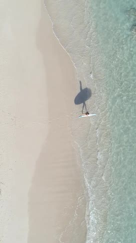 An aerial shot shows a person with a surfboard walking toward the ocean on a sandy beach next to clear shallow water