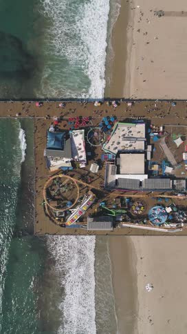 An aerial shot shows a bustling pier with a roller coaster other rides and numerous people alongside an ocean shoreline with waves breaking