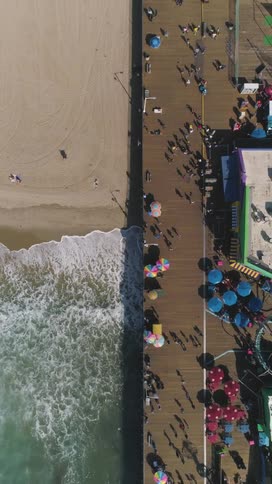 An aerial view shows a long wide pier with various colorful umbrellas and structures next to a sandy beach with crashing waves