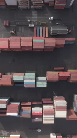 Aerial top view of a semitrailer truck driving next to rows of stacked shipping containers in varying colors mainly red