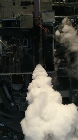 An aerial view shows thick white smoke billowing upwards from an industrial building with various pipes and structures