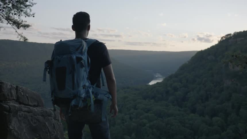 A man wearing a backpack approaches the overlook edge at dusk