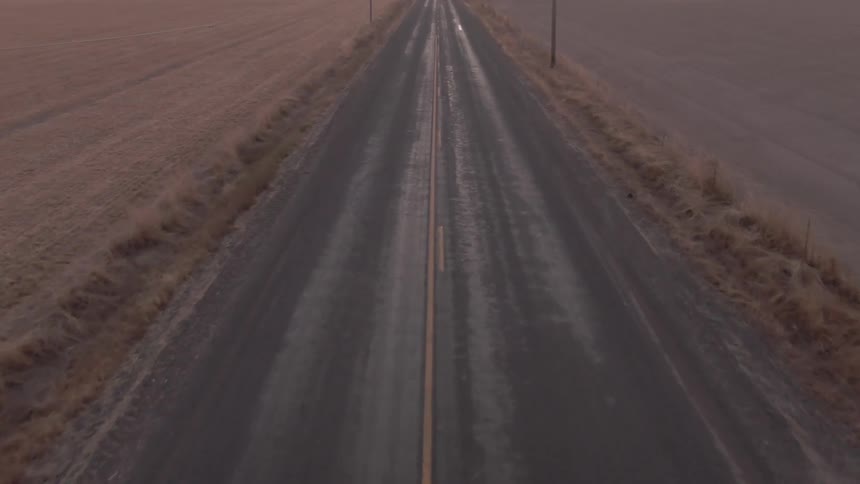 A straight empty road extends into the distance through flat brown fields under an overcast sky