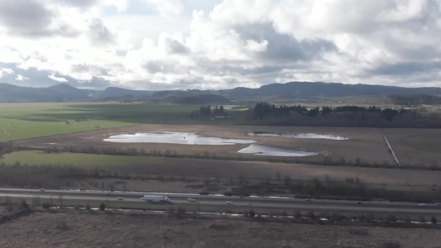 An aerial view shows a highway running through a landscape of fields and wetlands with mountains visible under a cloudy sky