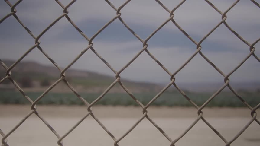 A blurred chainlink fence occupies the foreground looking out onto a green agricultural field a dirt path and distant hills under a cloudy sky