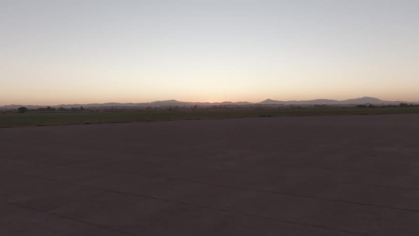 A vast empty field stretches out toward a distant mountain range under a clear early morning sky