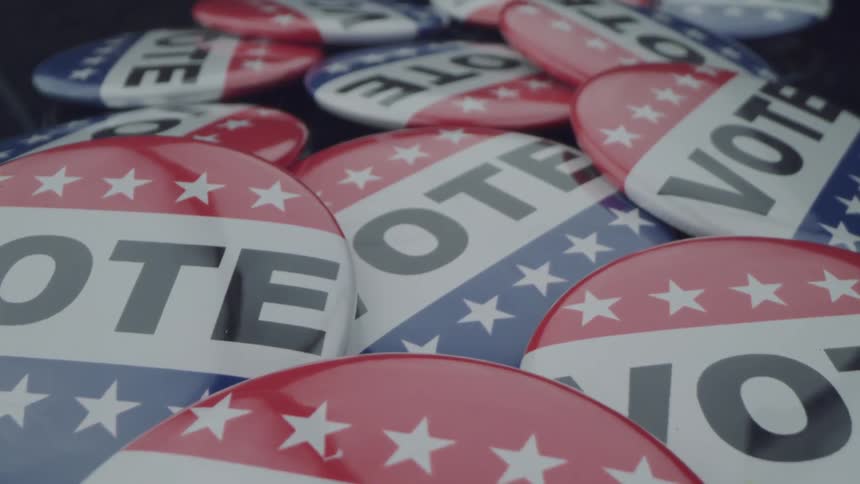 A collection of red white and blue buttons with the word vote printed on them is scattered across a surface