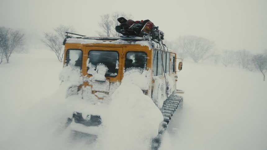 A yellow offroad vehicle drives through deep snow in a barren snowy landscape with sparse leafless trees