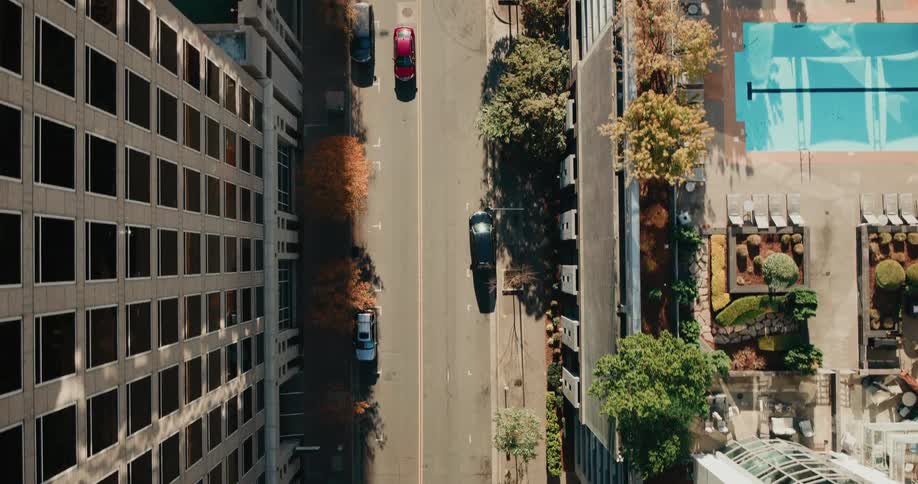An aerial view shows a city street and adjacent parking lot with scattered vehicles and shadows