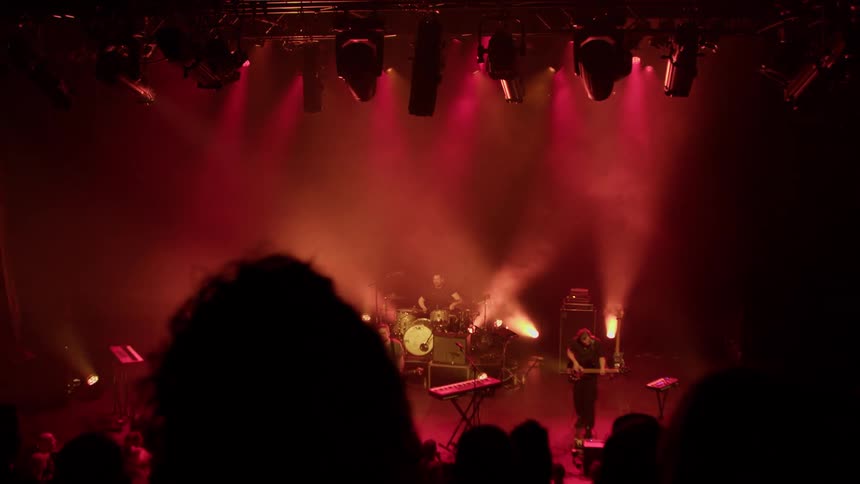 A band performs under intense red stage lighting with smoke effects as silhouettes of audience members watch in the foreground