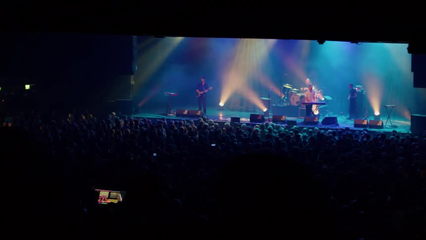 A band performs onstage in a large indoor venue under colorful lights with a cheering audience packed tightly in front