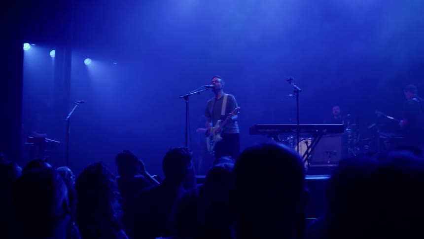 A man performs on stage during a concert playing the guitar and singing as people watch the atmosphere is bathed in blue light from the fixtures
