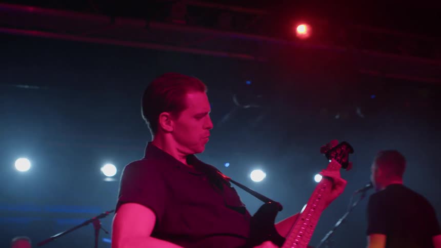 A musician plays an electric bass guitar on stage under red lighting during a live performance