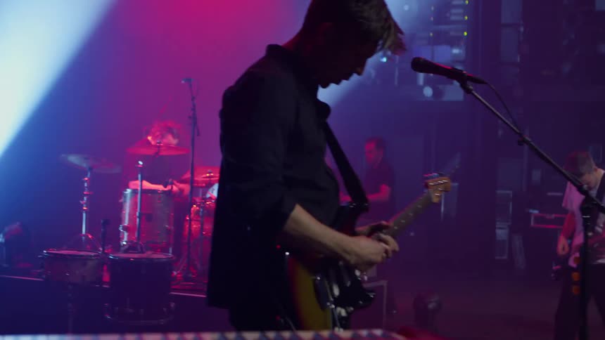 Male guitarist performs on a stage under lighting as part of band at a night concert