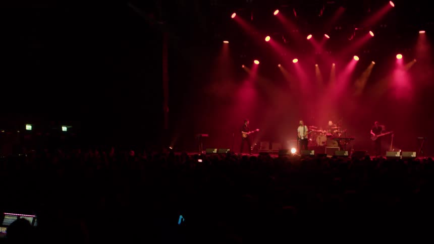 A live band performs on stage under dramatic red lighting in front of a large dark crowd