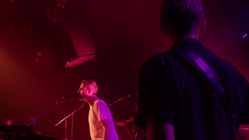 A band performs on stage under red and purple lights seen from behind a guitarist