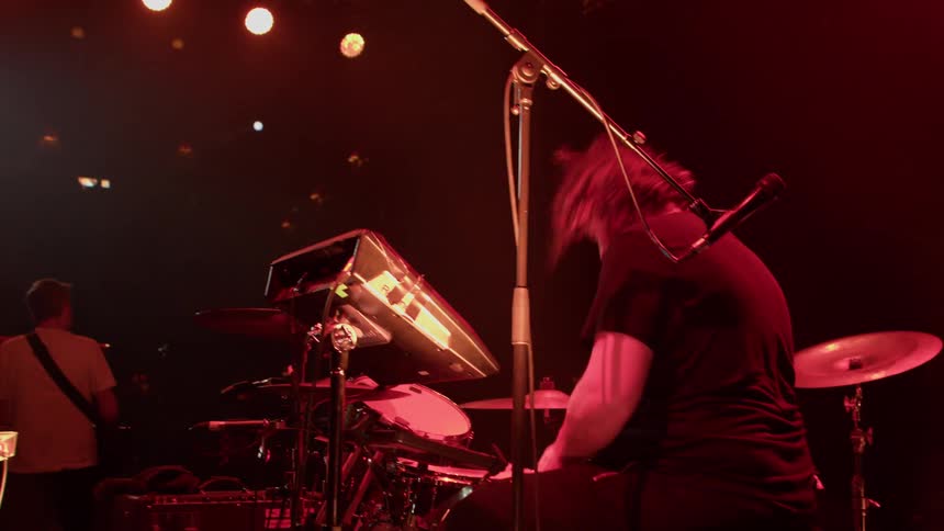 Male drummer plays on stage under red lighting as part of a band at a concert