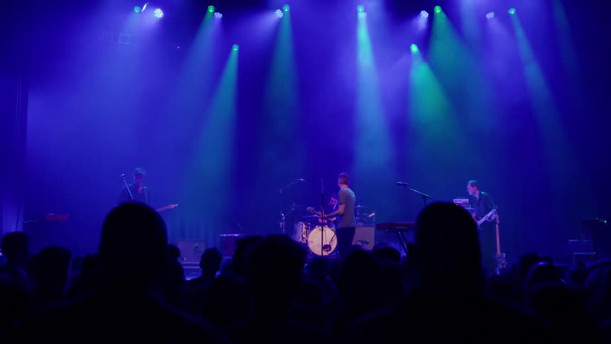 A live band performs on stage under blue and green lighting while a crowd watches in silhouette