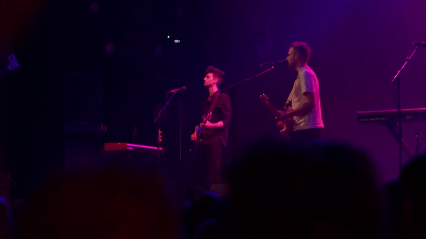 A live band performs under purple stage lighting during a concert in front of a crowd