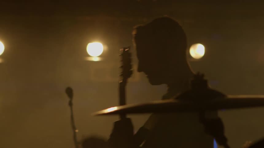 A musician performs onstage with a guitar under intense backlighting silhouetted against the stage glow before a large crowd
