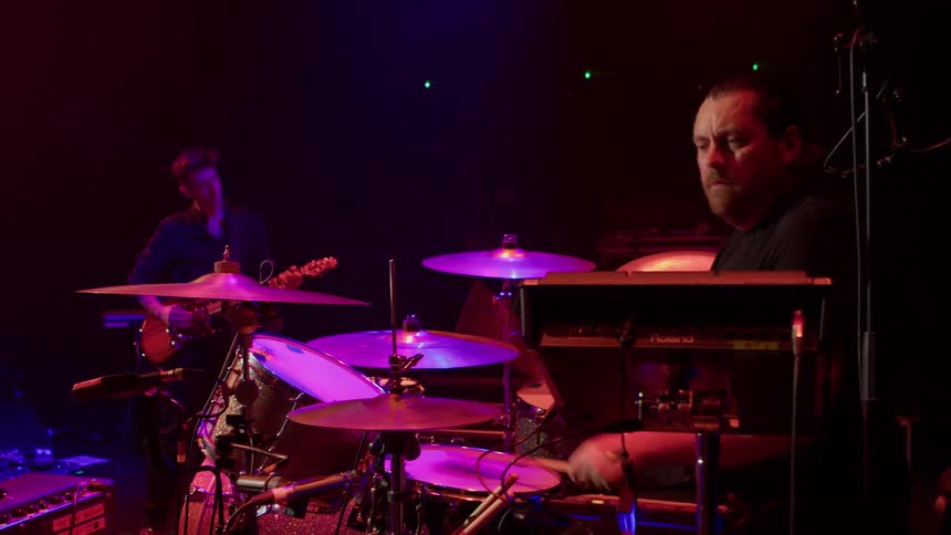 Male drummer and a guitarist perform on stage as part of a band at a concert at night
