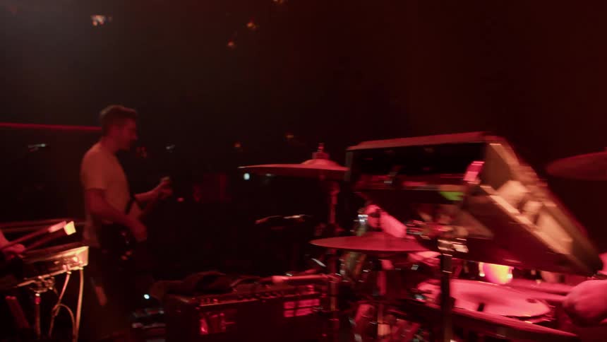 Two drummers face each other on a concert stage energetically playing their drums during the performance with a guitarist positioned in front of them