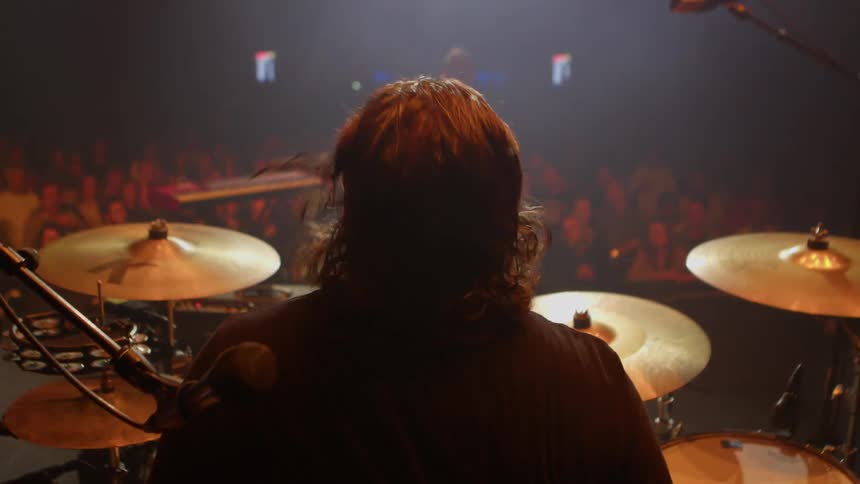 A drummer performs on stage with a band viewed from behind the drum kit as a crowd watches in the background