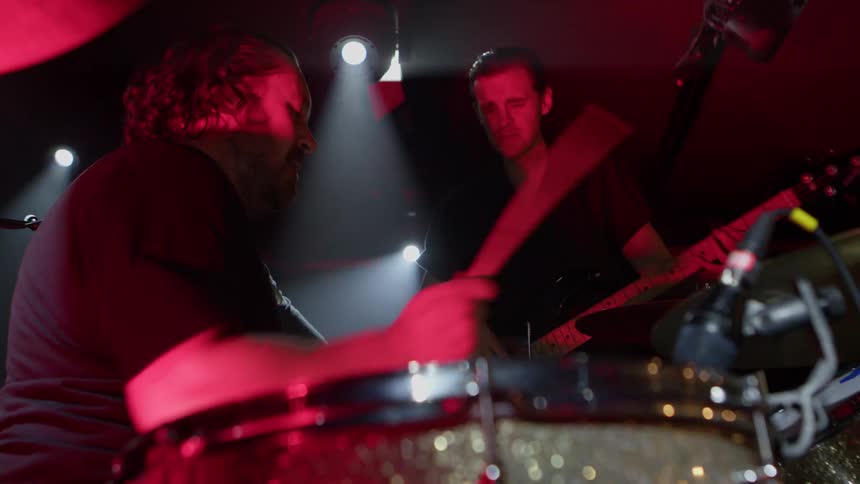 A drummer and a guitarist perform onstage under dramatic lighting surrounded by amplifiers and stage equipment