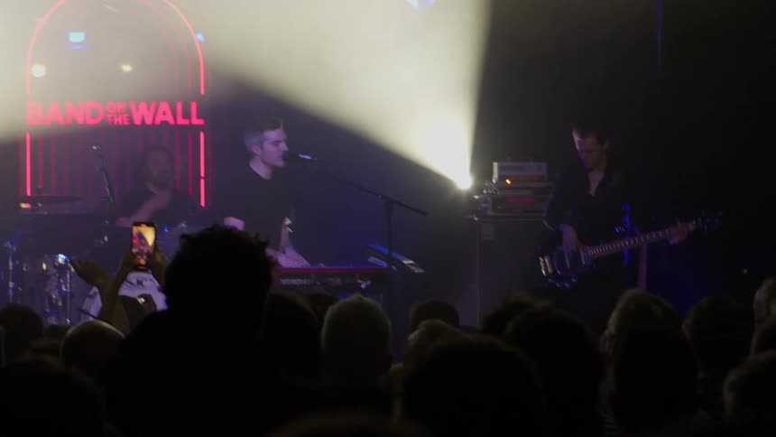 A man singing and playing a guitar alongside other guitarists and band members during a musical concert as the crowd cheers and watches