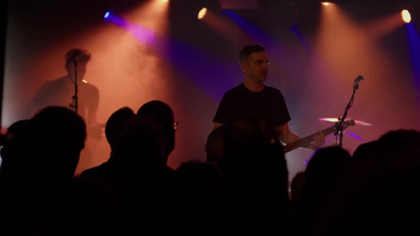 A band performs onstage under colorful lighting with a packed crowd in front of the stage
