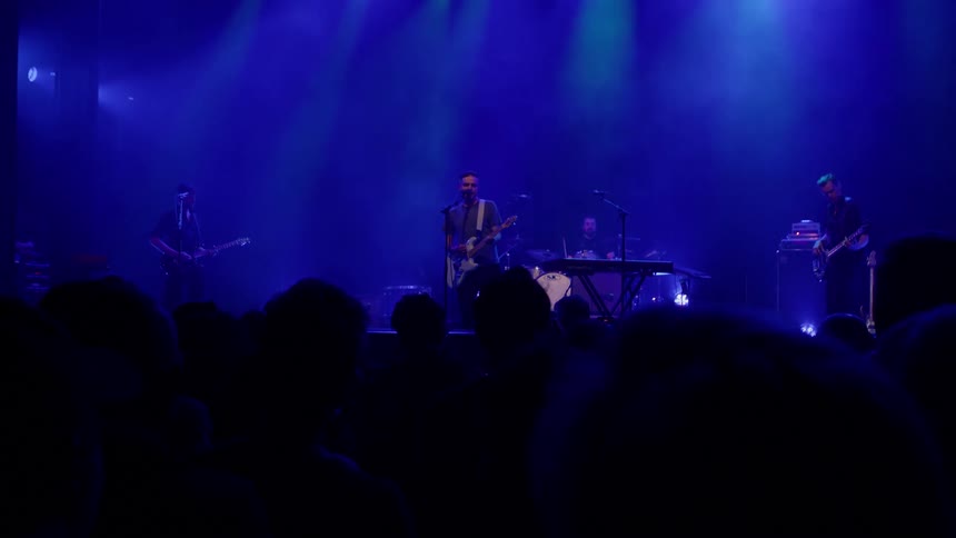 A male band performs on stage under lighting in front of a crowd at night