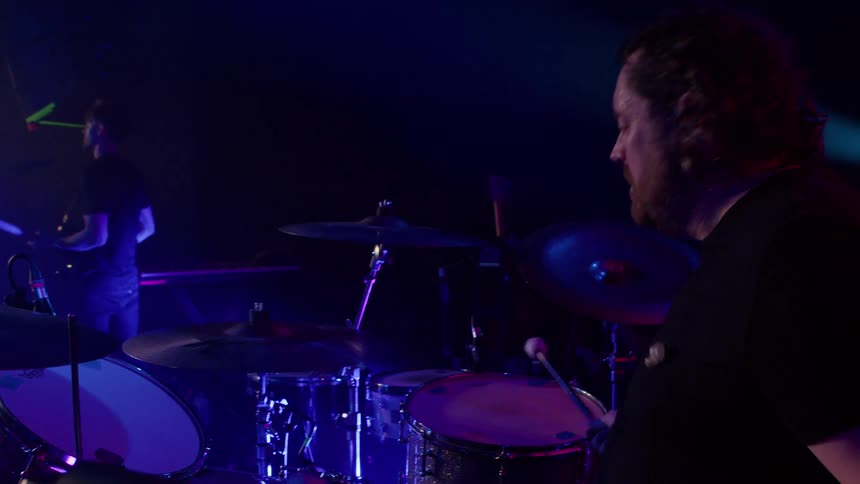 A man plays in a band with another man in front playing a guitar during a music concert on stage at night