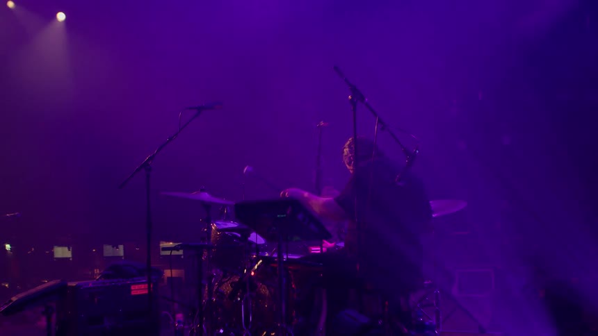 A band performs live on stage in moody purple lighting with a focus on the drummer and guitarist