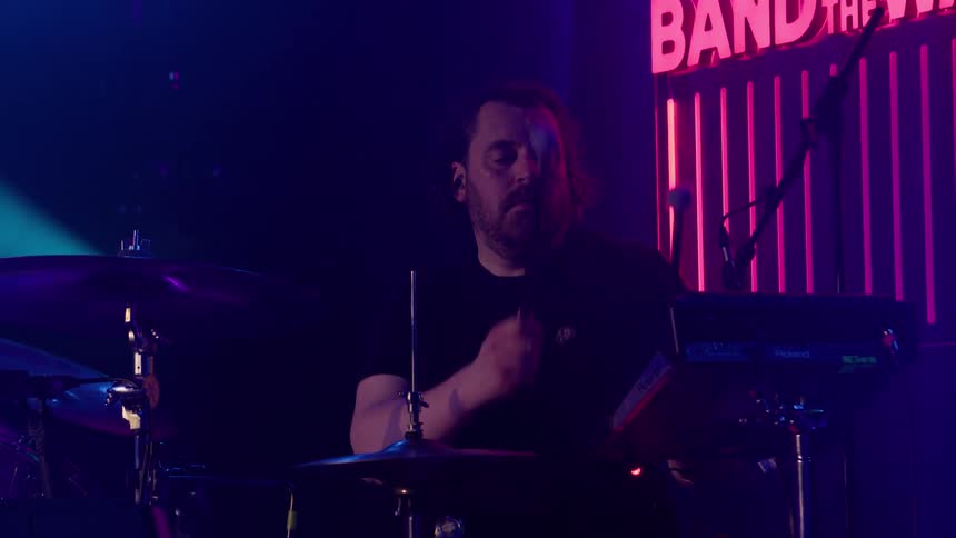 A drummer plays with concentration on a dark stage illuminated by blue and pink neon lights