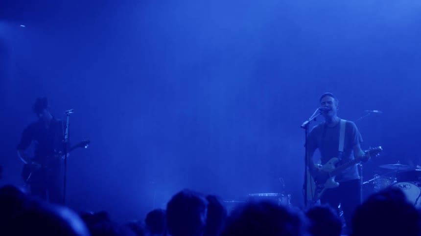 A man singing and playing guitar during a concert accompanied by other musicians playing drums guitar and bass