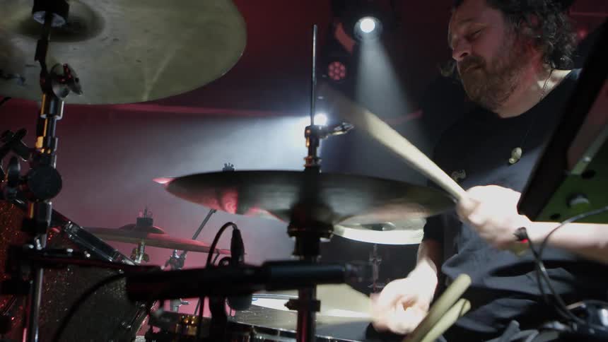 A drummer intensely plays his kit under dramatic stage lighting during a live performance